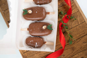  Chocolate-covered brownie popsicles with decorative hazelnuts and green leaves, placed in a white box with red ribbon.