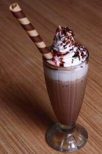  A tall glass of chocolate brownie milkshake topped with whipped cream, sprinkles, chocolate syrup, and a rolled wafer stick.