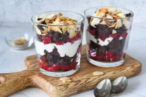 A layered dessert in a glass featuring crumbled brownies, whipped cream, fresh red fruits, and toasted almond slices.