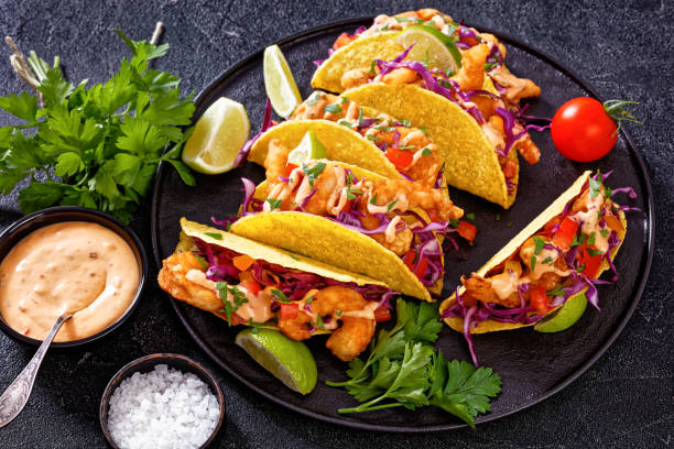 Bang Bang Shrimp tacos served in crispy yellow tortillas with red cabbage, tomatoes, and lime wedges.