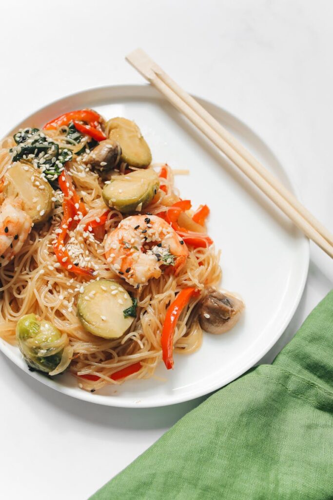 Shrimp stir-fry with rice noodles, Brussels sprouts, red peppers, mushrooms, and sesame seeds on a white plate with chopsticks.