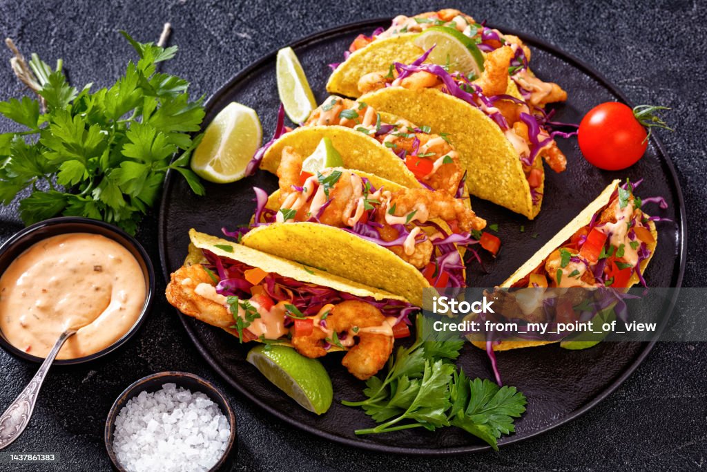 Crispy shrimp tacos drizzled with Bang Bang Sauce, garnished with fresh vegetables and lime wedges on a black plate"