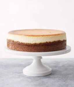 A perfectly baked classic cheesecake with a golden top and smooth layers, sitting on a white cake stand.