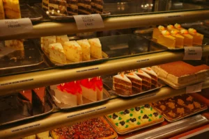 A Cheesecake Factory bakery display case filled with a variety of desserts, including cakes, cheesecakes, brownies, and vegan options, neatly labeled and arranged on multiple shelves.