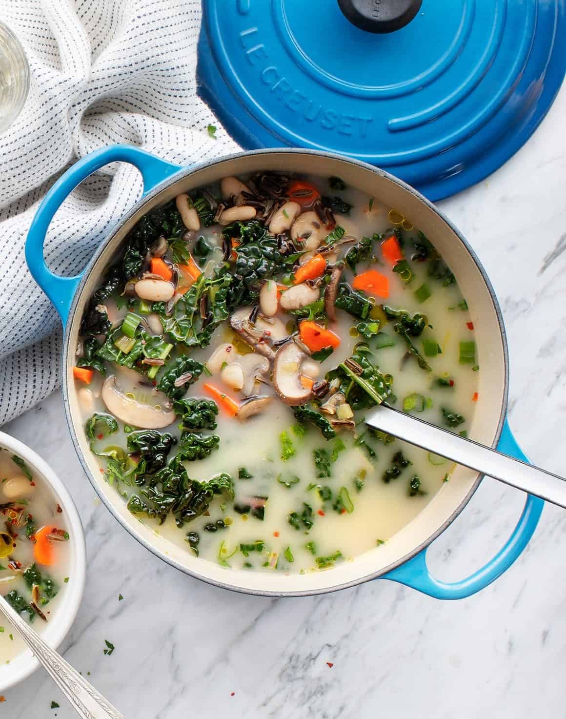 Healthy vegetable and white bean soup in a blue pot, featuring kale, carrots, and mushrooms